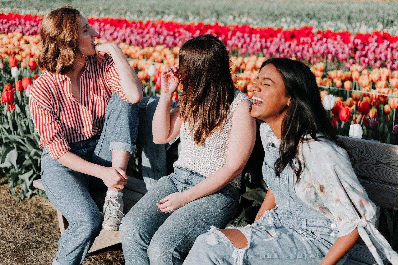 Three girls sitting down laughing in front of field of flowers